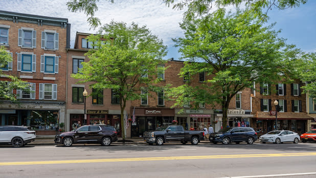 A quaint street scene in Skaneateles with brick buildings housing storefronts and green trees lining the sidewalk. Parked cars and a few pedestrians are visible under a partly cloudy sky.