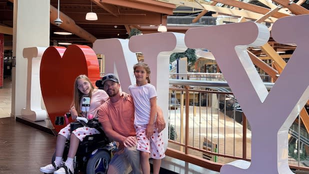 A man and two children, one using a wheelchair, smile in front of a large "I LOVE NY" sculpture at the Destiny USA mall.