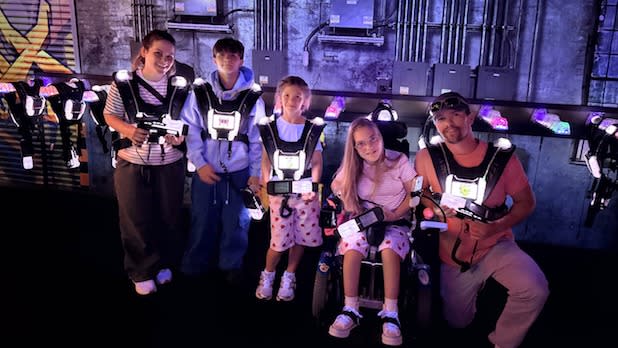 A group of five people in a dimly lit room, all wearing futuristic, glowing vests for a laser tag game at Apex Entertainment at Destiny USA.