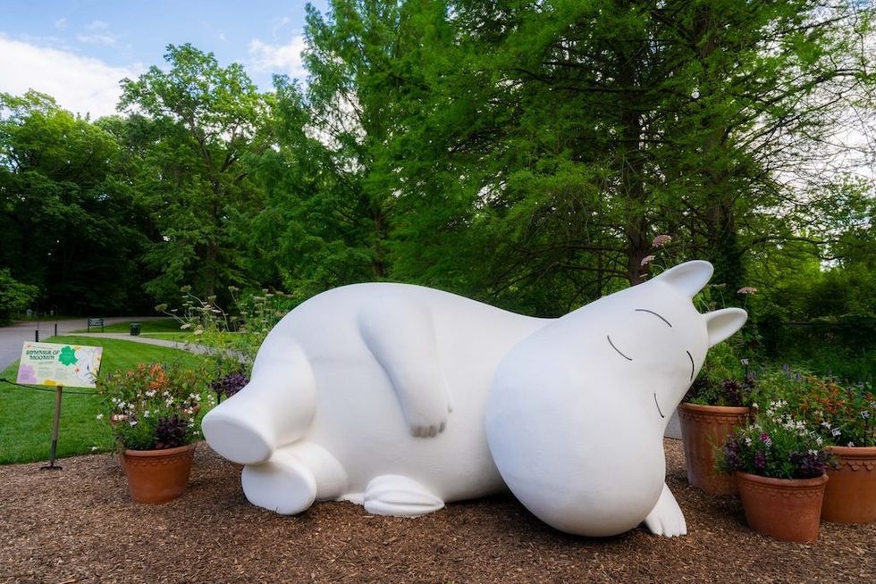 Large white Moomin sculpture sleeping in a garden with lush greenery.