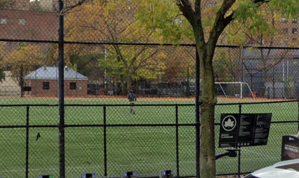 Playground with green turf behind a chain-link fence, with a building, soccer goal, and trees visible.