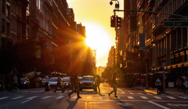 A city street at sunset with people crossing the road, cars stopped at a traffic light, and tall buildings lining both sides—capturing the vibrant life and energy of the Manhattan real estate market bathed in warm golden sunlight.
