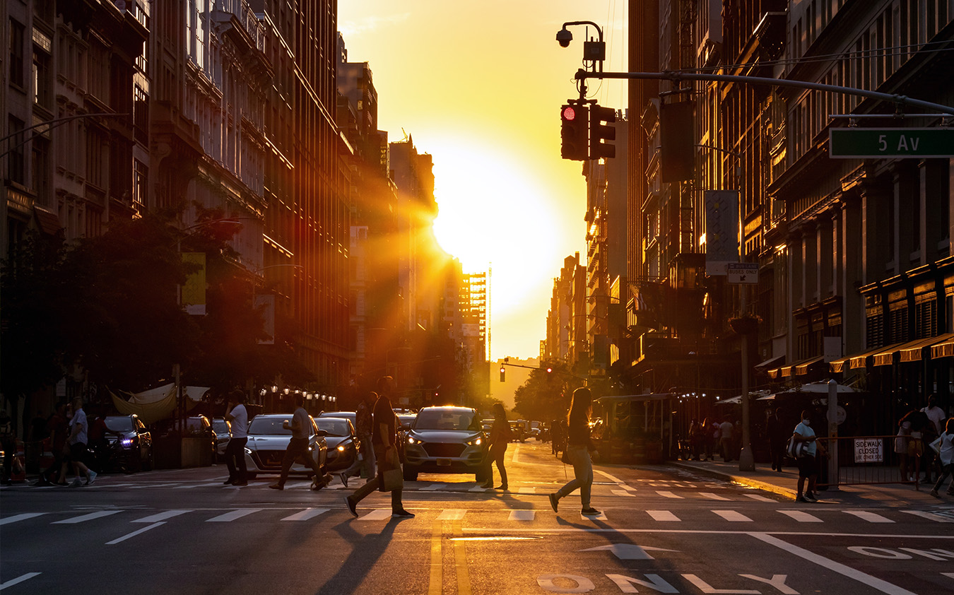 A city street at sunset with people crossing the road, cars stopped at a traffic light, and tall buildings lining both sides—capturing the vibrant life and energy of the Manhattan real estate market bathed in warm golden sunlight.