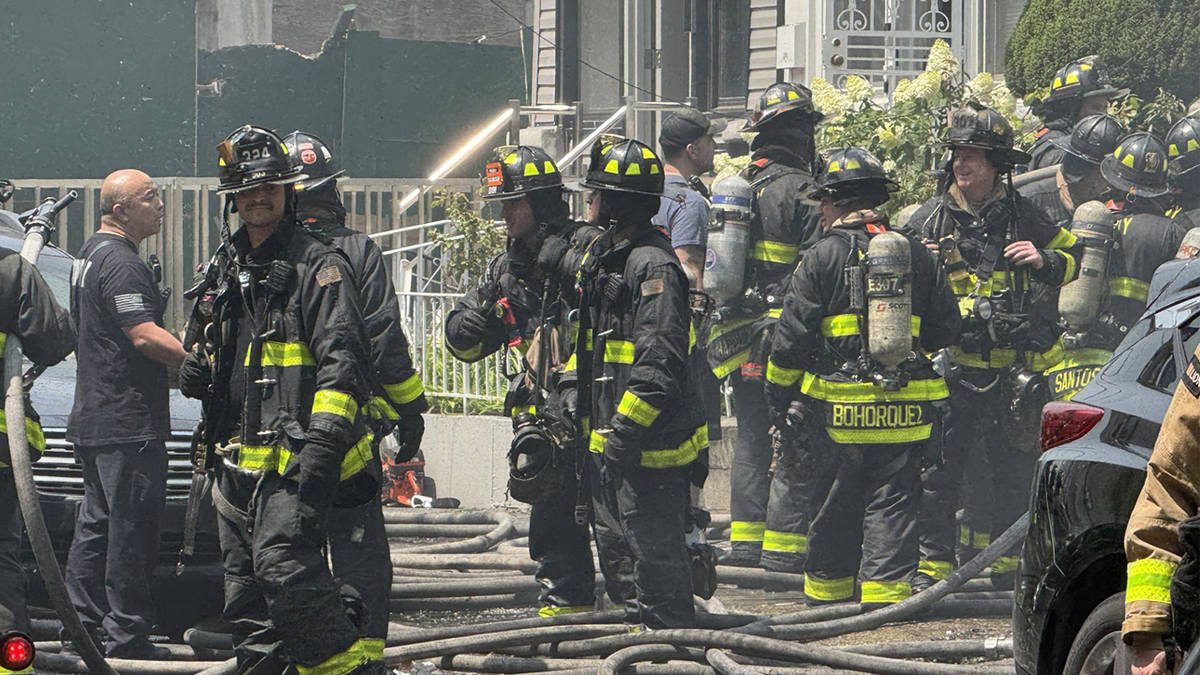 Firefighters gather outside a residential building as crews continue battling a large structure fire with hoses stretched across the street.
