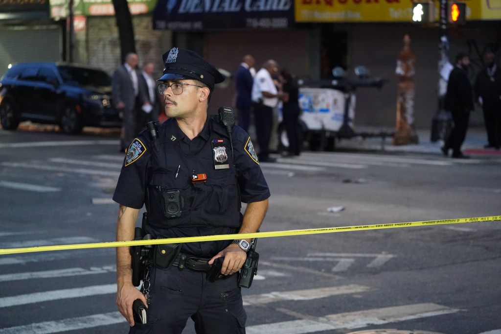 A police officer with a mustache and glasses stands at a crime scene blocked off by yellow tape.