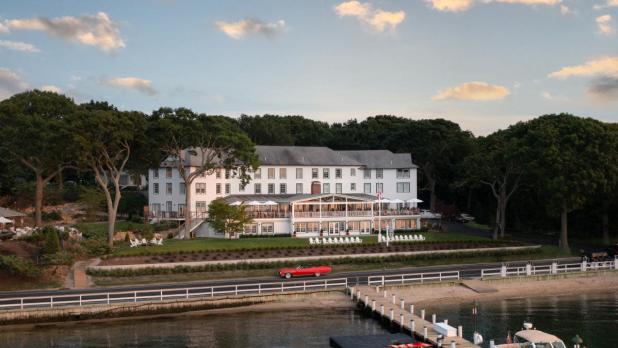 Aerial view of the Pridwin Hotel & Cottages, a large, elegant white building surrounded by lush trees. A calm waterfront with docks and boats is in the foreground. Evening sky sets a serene mood.