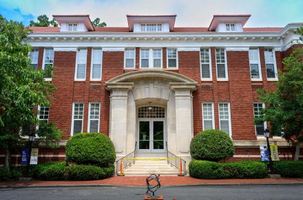 A photo of the exterior of Queens Hall's main entrance. Limestone stairs lead up to two columns with lush landscaping on either side of the staircase.