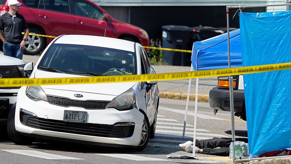 A white car with multiple bullet holes in the windshield stopped behind yellow police tape.