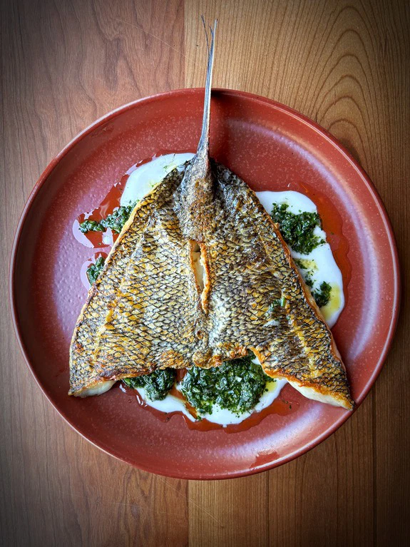 Grilled fish served on a red plate with green sauce and creamy elements on a wooden table surface.
