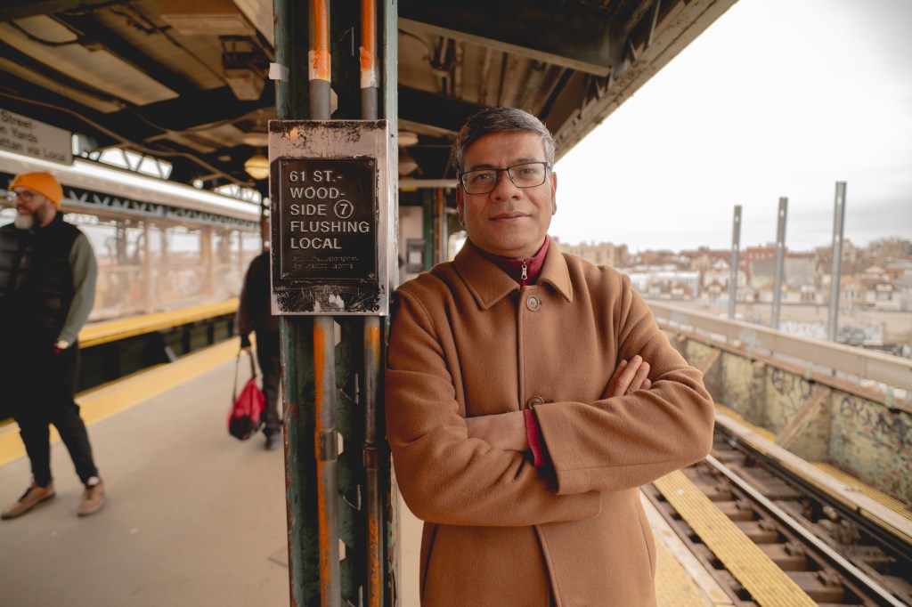 Shamsul Haque stands with arms crossed on a subway platform next to a sign for 61 St. - Woodside and the 7 Flushing Local train.