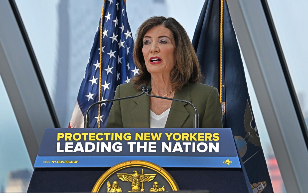 Governor Kathy Hochul speaking at a podium with the text "PROTECTING NEW YORKERS LEADING THE NATION" and American and New York State flags behind her.