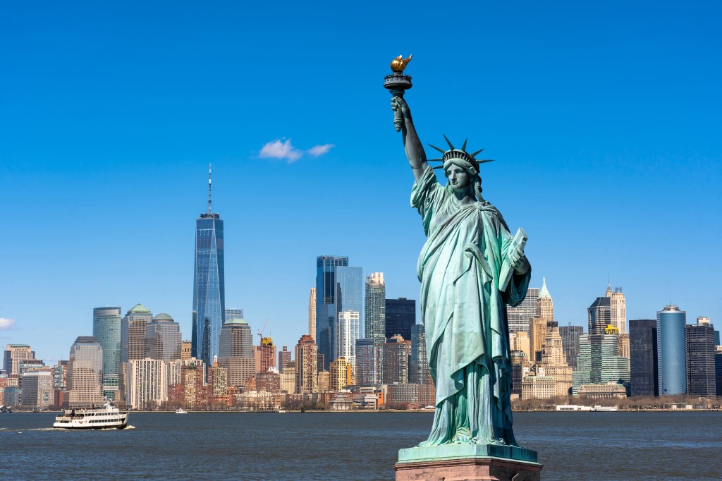 The Statue of Liberty in the foreground with the Lower Manhattan skyline, including One World Trade Center, and the Hudson River with a ferry in the background.
