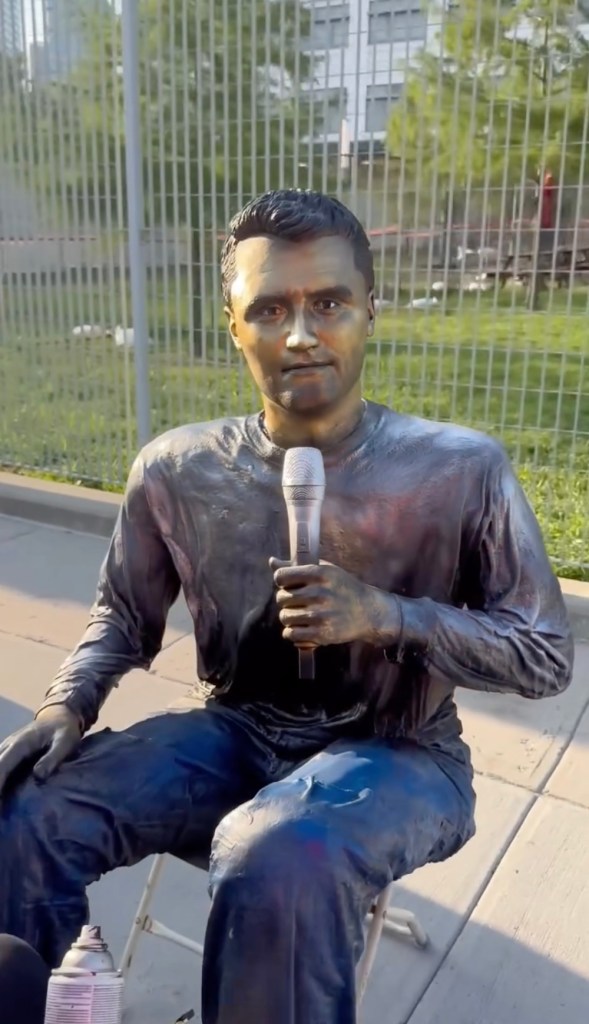 A person painted to look like a bronze statue of Charlie Kirk holding a microphone.