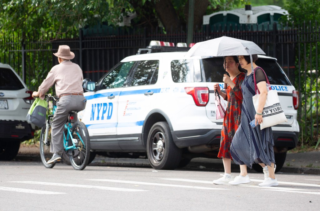 Two women with an umbrella cross a street while a man on a bicycle passes by an NYPD vehicle.