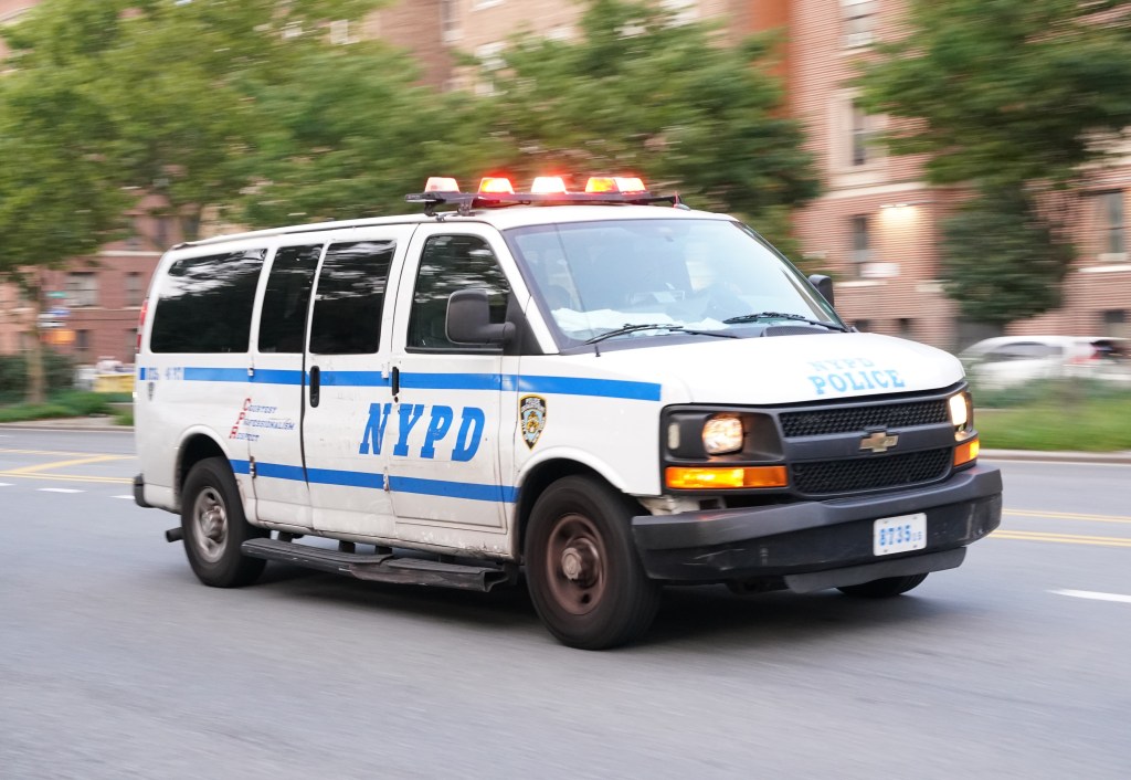 A white NYPD police van with blue stripes, emergency lights, and "NYPD" written on the side, driving on a street.