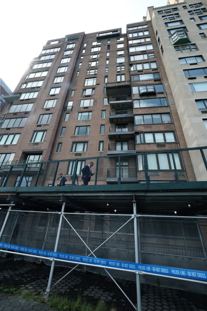 A tall brick building with police line tape across the bottom, with two men standing on a balcony overlooking the street.