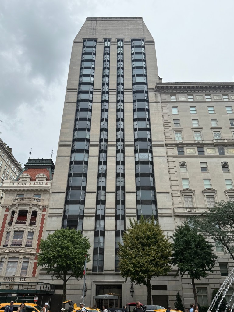 A tall modern building with a concrete facade and large glass windows, flanked by two trees and situated near older architectural structures, under a cloudy sky.