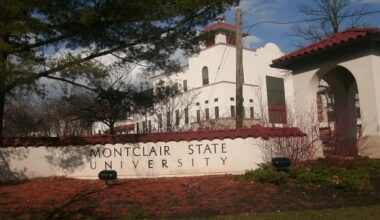 White stucco entrance and tower at Montclair State University, with the university name displayed on a low wall in the foreground and trees surrounding the building.