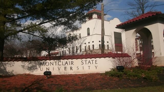 White stucco entrance and tower at Montclair State University, with the university name displayed on a low wall in the foreground and trees surrounding the building.