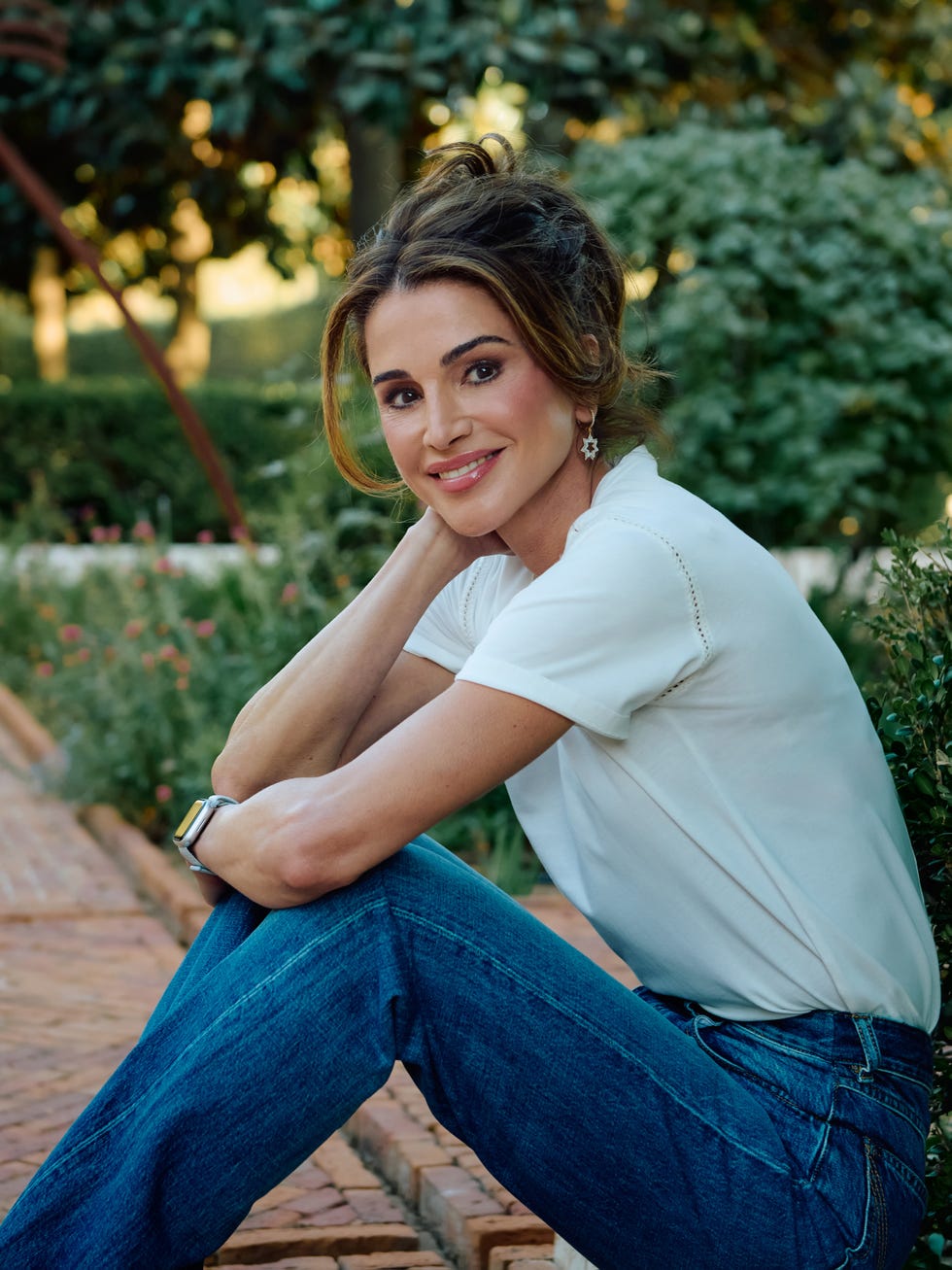 A woman with long hair sits outdoors, smiling and resting her chin on her hand, wearing a white top and blue jeans.