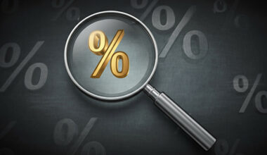 Image of an interest rate sign with a magnifying glass hovering over it. Black and gray background with interest rates in gray on it.