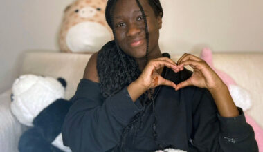 A girl with a black shirt and long black braids makes a heart cymbol with two hands. She is sitting on a bed with stuffed animals around her.