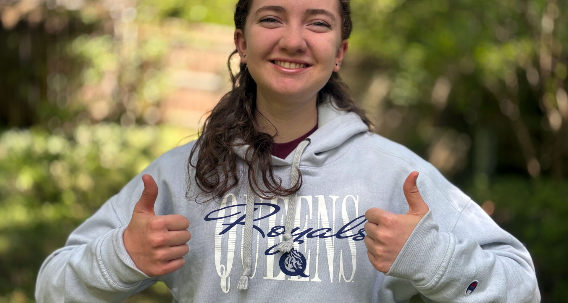 Girl wearing a gray Queens Royals sweatshit gives two thumbs up on a nature trail.