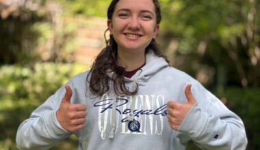 Girl wearing a gray Queens Royals sweatshit gives two thumbs up on a nature trail.