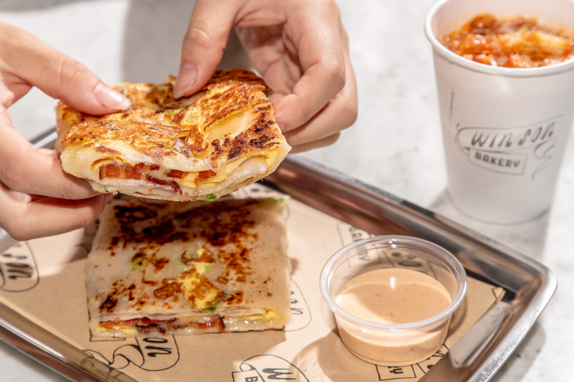 Two hands holding a folded scallion pancake egg and bacon sandwich.
