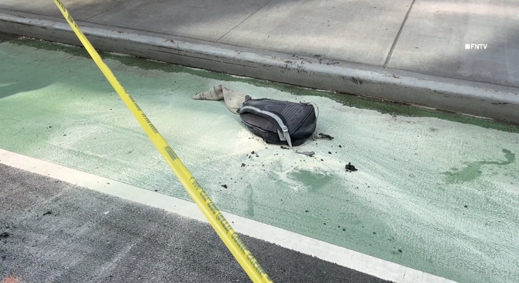 Yellow caution tape blocks off a section of green pavement with a dark backpack, a piece of clothing, and white powder on it.