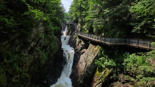 A cascading waterfall flows between steep, lush green cliffs, with a wooden walkway on the right side at High Falls Gorge in Wilmington, NY.