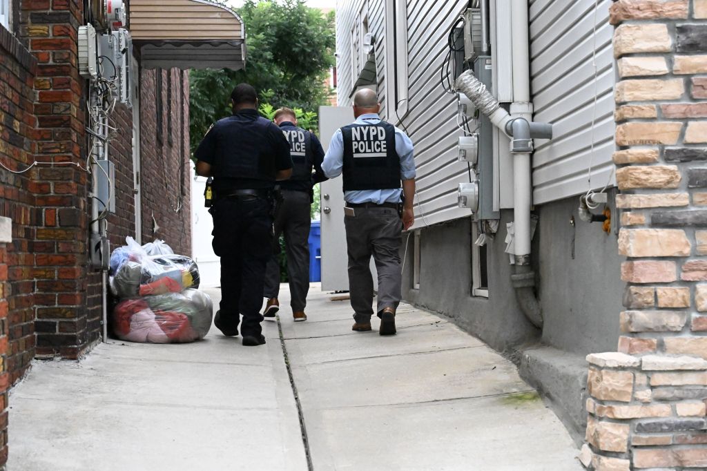 Three NYPD officers walk down an alleyway next to a building with siding and exposed pipes.