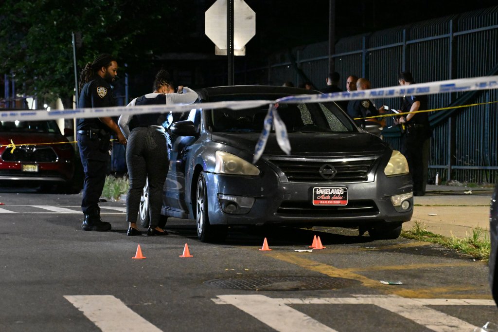 Police officers and a woman wearing a bulletproof vest examine a car at a crime scene.