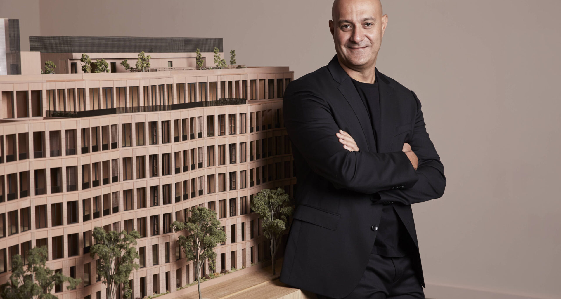 A man in a black suit stands with arms crossed next to a detailed architectural model of a multi-story building with trees.