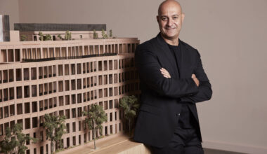 A man in a black suit stands with arms crossed next to a detailed architectural model of a multi-story building with trees.