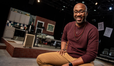 A man sits and poses for a photo. In the background is the set of a theatre production, including an apartment set.