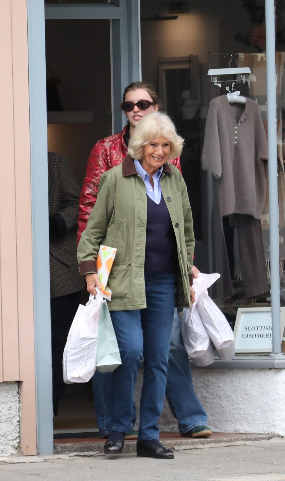 Two women exiting a store, one carrying shopping bags and smiling, with another in the background wearing sunglasses.