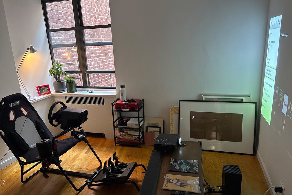 A cozy gaming setup featuring a racing simulator chair, projector, and a mix of books and decor in a bright room.