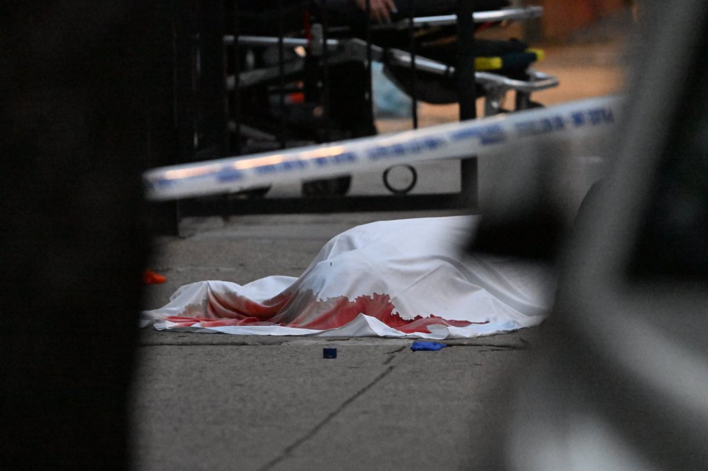 A white sheet covers a body on the ground, stained with blood, with crime scene tape visible in the foreground.