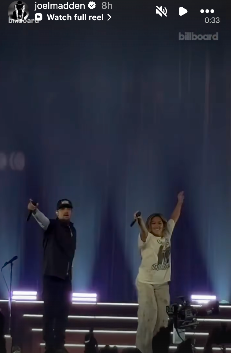 Two performers on stage, one holding a microphone and the other raising their hand in excitement, with bright lights in the background. Two performers on stage, one holding a microphone and the other raising their hand in excitement, with bright lights in the background.