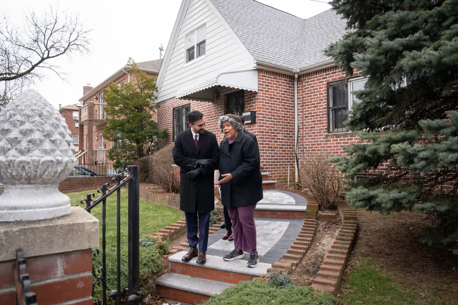 Two people walk together on a brick pathway beside a brick house with white trim and a slate roof. Evergreen shrubs and bare trees line the sides of the walkway.