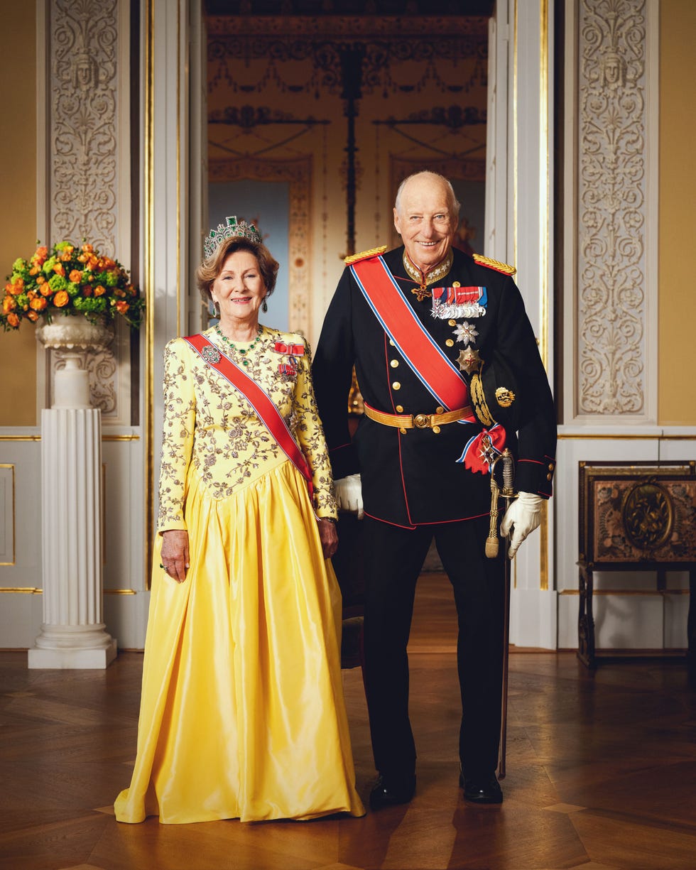 A royal couple dressed in formal attire, posing in an ornate room with floral decorations.