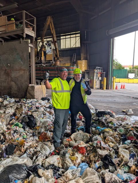 Two workers in high-visibility vests standing on a large pile of trash in a processing facility.