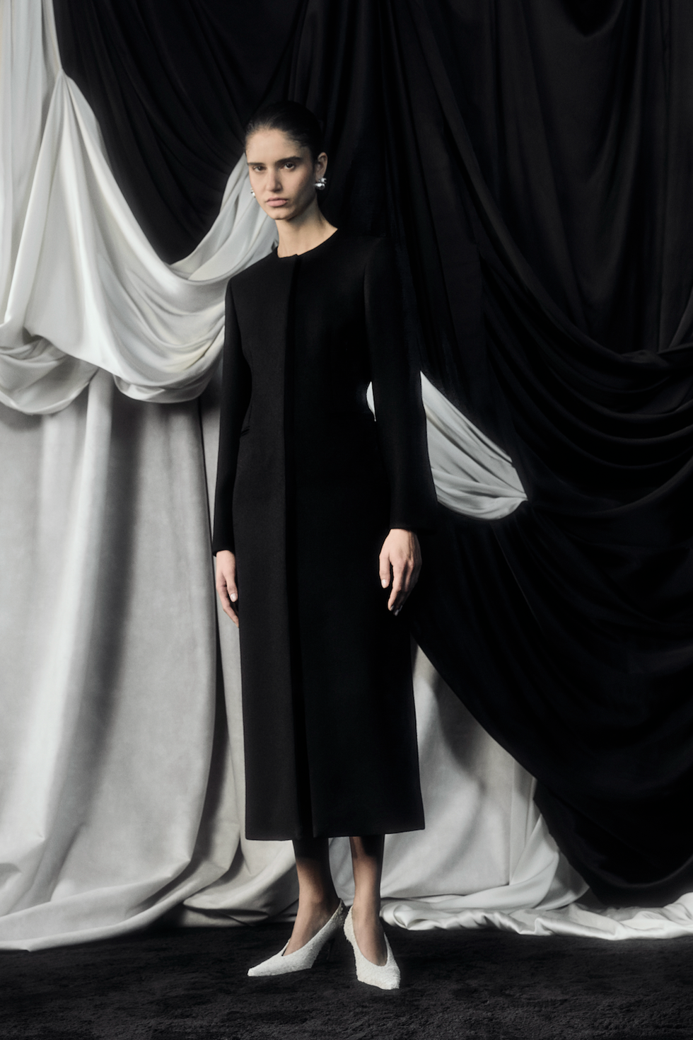 A model wearing a long black coat stands against draped fabric in black and white, complemented by white heels.