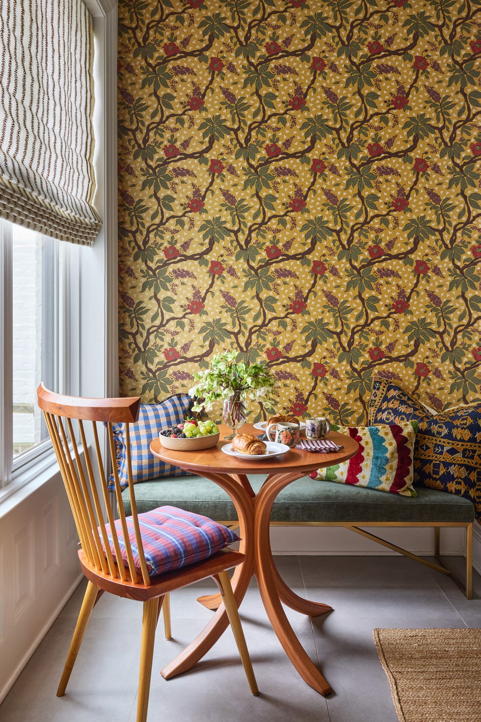 Cozy breakfast nook with floral wallpaper, a wooden table, assorted fruits, and pastries on a patterned cushion seating.