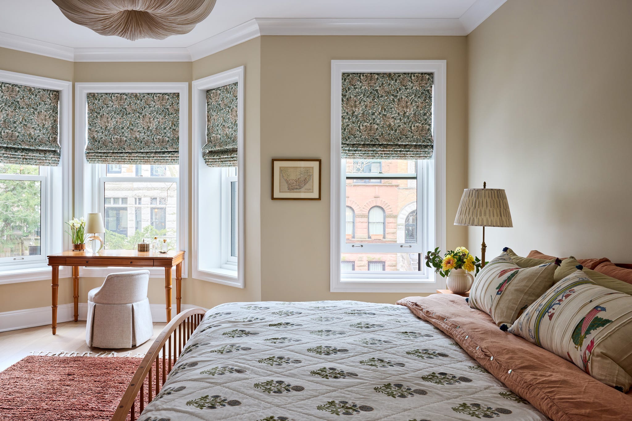 Cozy bedroom featuring warm colors, floral accents, a wood bed frame, and a small desk by large windows with patterned shades.