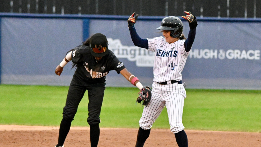 A Los Angeles Queens baseball player dressed in a black jersey tags a New York Heights player in a white jersey with blue stripes. The Heights player is celebrating because she is already on-base and safe.