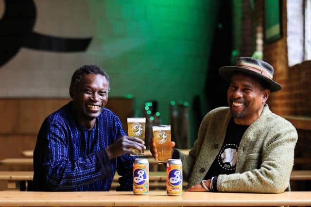 African brewer Pierre Thiam Yolélé and Brooklyn Brewery’s Garrett Oliversmiling and holding up glasses of beer next to two cans of Brooklyn Brewery beer on a table.