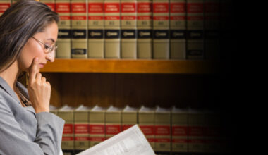 A woman in a gray suit and glasses reads a book, standing in front of shelves filled with legal books.