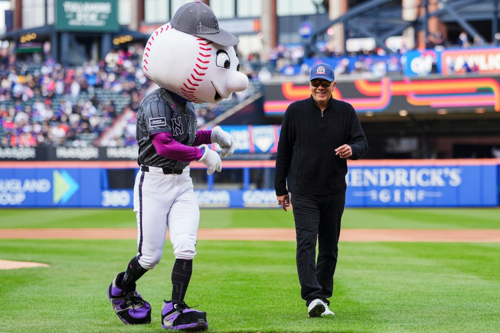 Mr. Met mascot walking with a man in a black sweater and baseball cap on a baseball field.